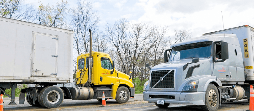 Yellow semi truck and grey semi truck parked in paved training yard. Cabs facing each other.