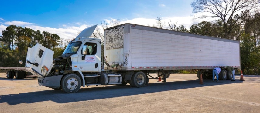 A CDL student inspects the rear trailer tires of a semi-truck with its hood open