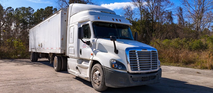 A white Class A semi-truck with a trailer parked on pavement, with trees and a blue sky in the background