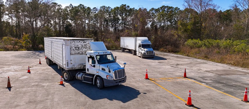 Two white semi trucks with trailers practice maneuvers on a CDL training range marked with orange cones and yellow painted lines, surrounded by pine trees.